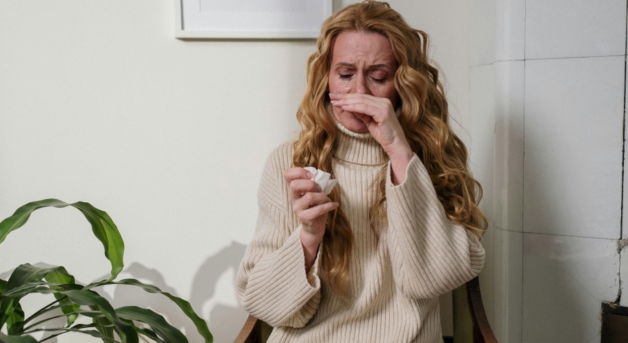 Woman in a beige sweater holding tissues and wiping her nose.