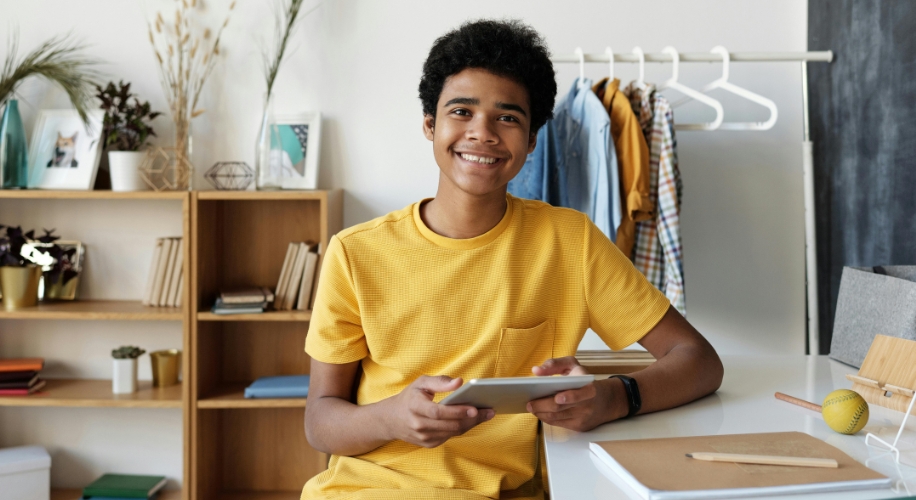 Teenager wearing a yellow shirt, holding a tablet and smiling while sitting at a desk.