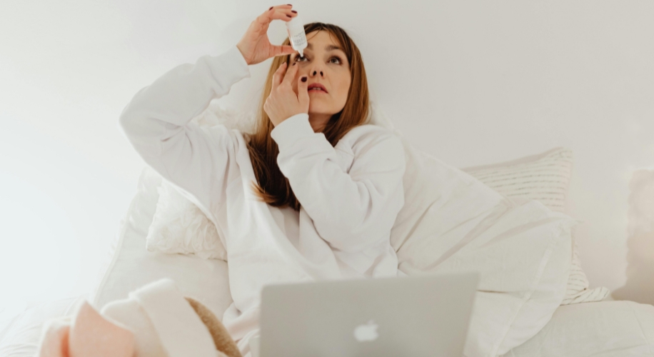 Woman using eye drops while sitting on a bed.