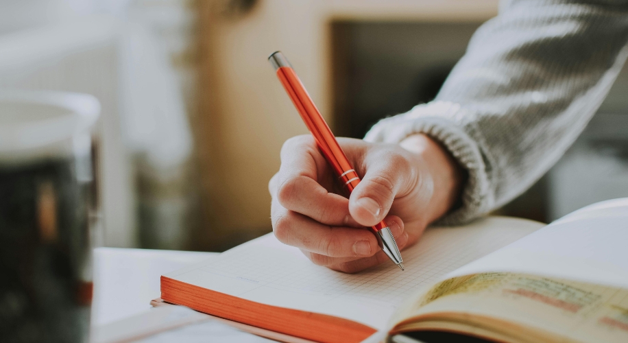 Hand holding a red pen, writing in an open notebook with grid lines.