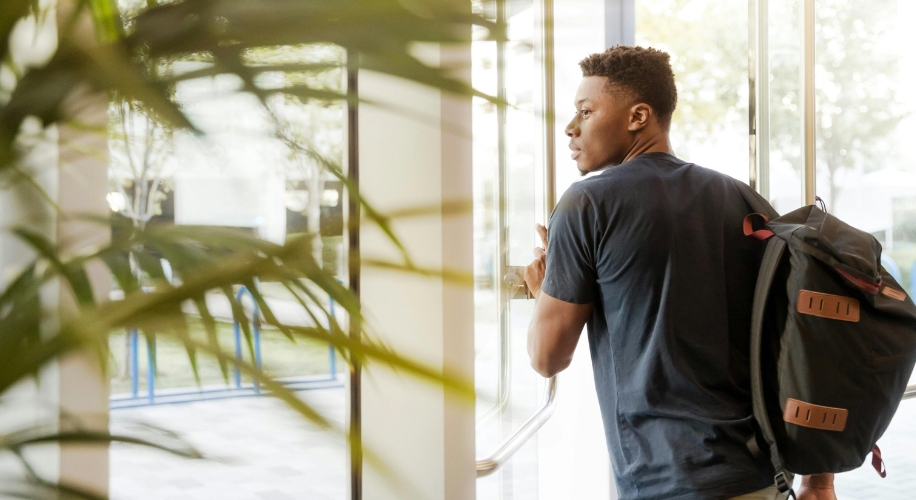 Person with a black backpack and blue shirt opening a glass door.