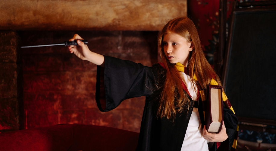 Child with a wand and book wearing a black robe and striped scarf in black, yellow, and red colors.