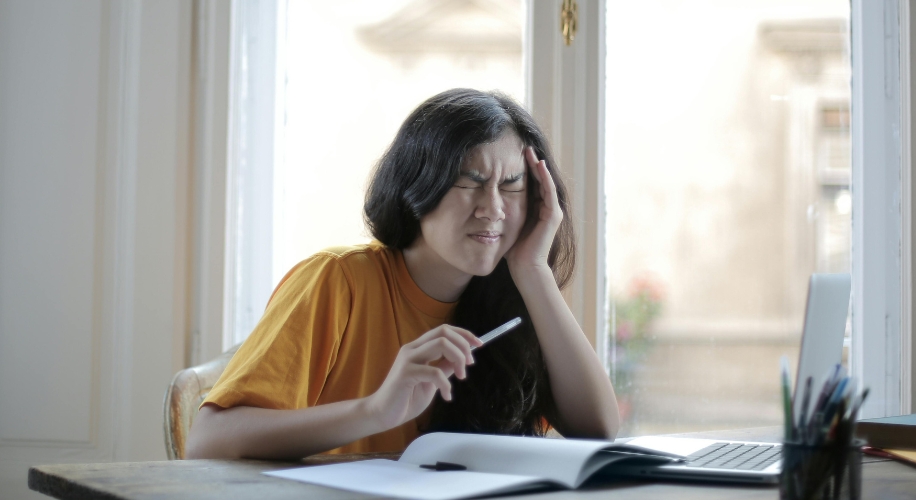 Person in a yellow shirt holding a pen, grimacing with their hand on their head, sitting in front of a laptop.