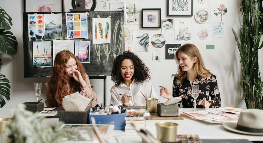 Three women enjoying a painting session with various art supplies on a table, surrounded by paintings.