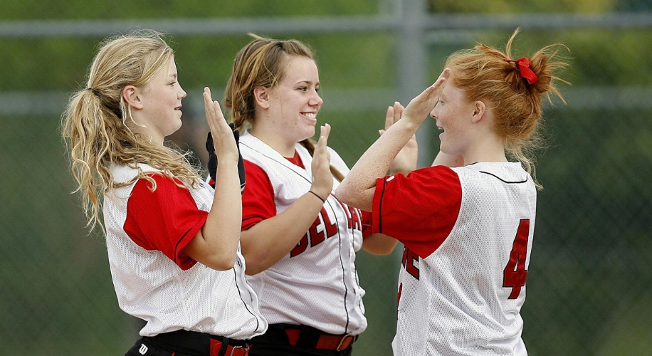 Three female softball players in white and red uniforms high-fiving each other with smiles on their faces.