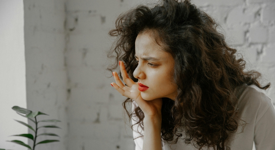 Woman with curly brown hair, red lipstick, and an orange manicure, looking confused or curious.