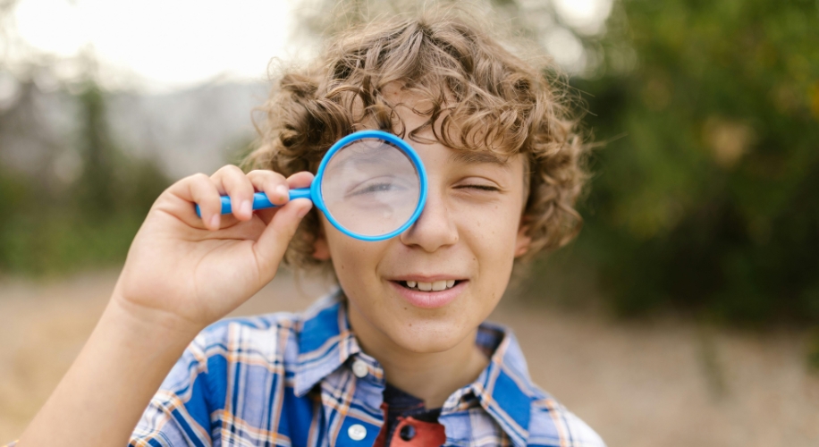 Boy holding a blue magnifying glass, examining closely with one eye through the lens, wearing a plaid shirt.