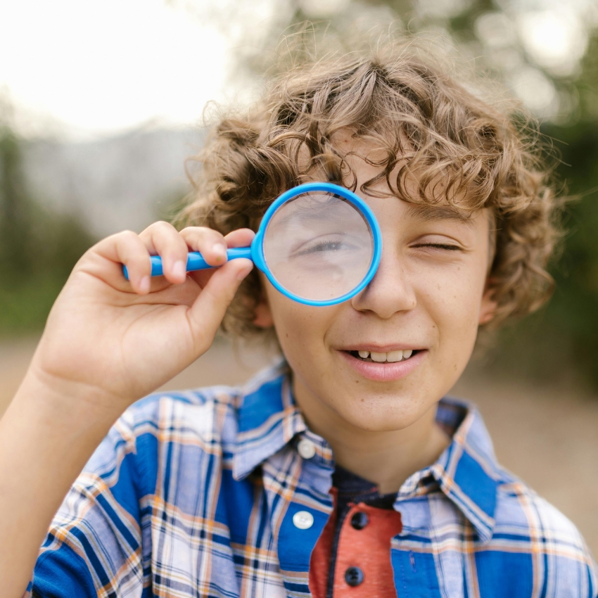 Child holding a blue magnifying glass up to one eye, wearing a blue plaid shirt.