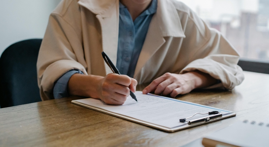 Person in a beige coat writing on a form attached to a clipboard with a pen.