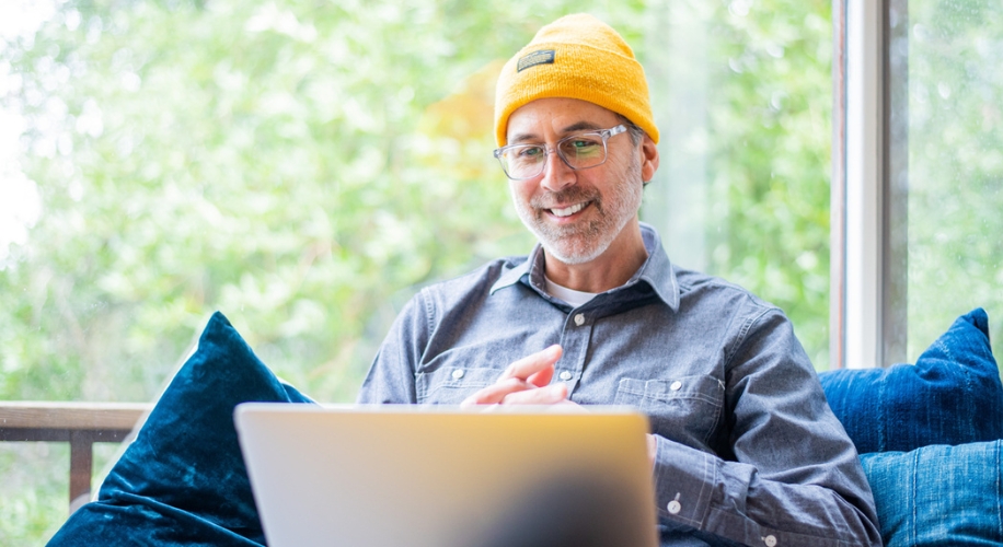 Man in yellow beanie and glasses using a laptop, sitting on a couch with blue pillows.