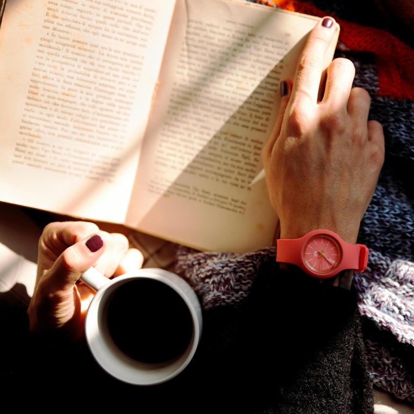 Person wearing a pink wristwatch and holding an open book and a coffee cup.