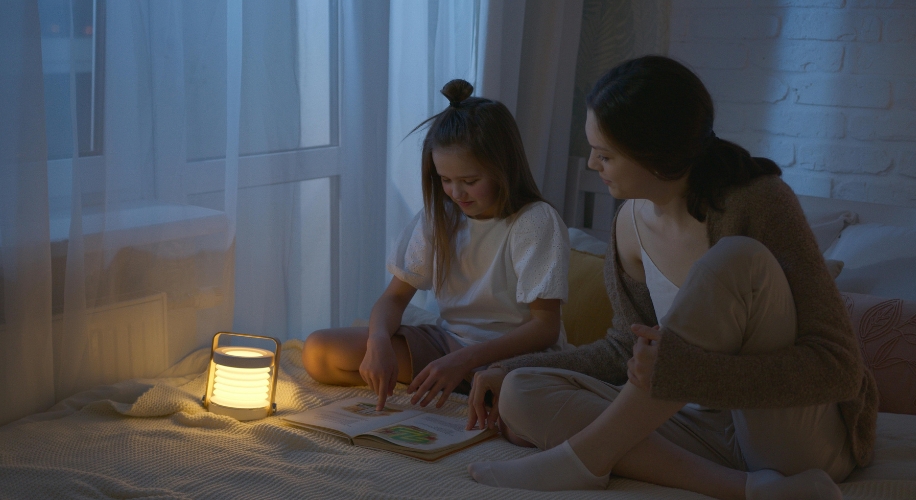 Child and adult reading a book by a soft, glowing lantern on a bed.