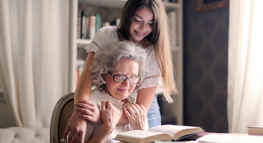 Elderly woman reading a book while a young woman gently hugs her from behind.