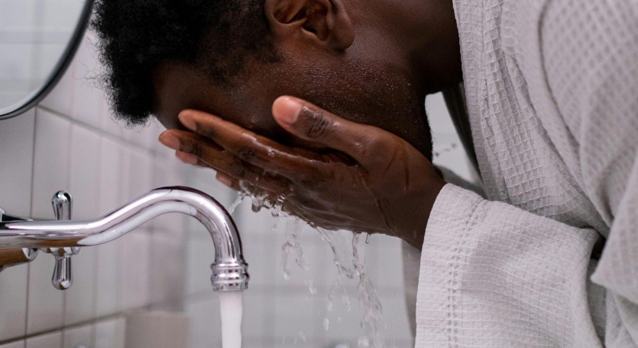 Person washing face with water from a modern faucet, wearing a white waffle-textured bathrobe.