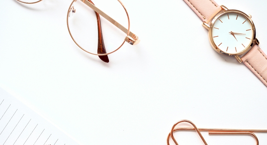 Round gold eyeglasses, a wristwatch with a white face and rose gold strap, and gold paperclips.