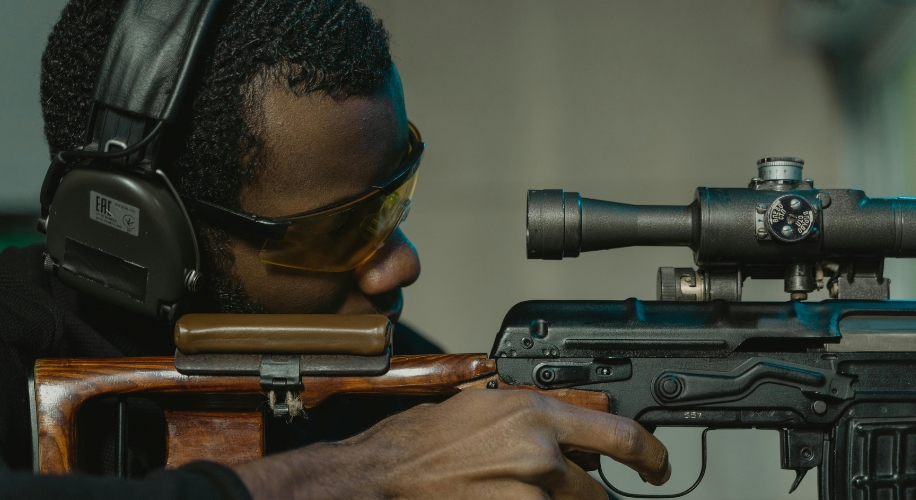 Person aiming a rifle with a scope, wearing ear protection and yellow-tinted safety glasses.