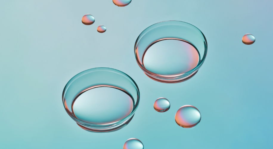 Transparent contact lenses with a hint of rainbow reflection surrounded by water droplets against a light blue backdrop.