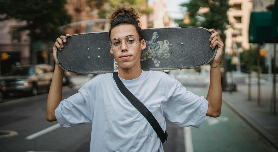 Person holding a skateboard behind their neck, wearing glasses and a white t-shirt with a black strap.