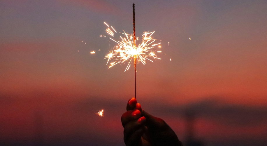 Hand holding a lit sparkler against a sunset sky.