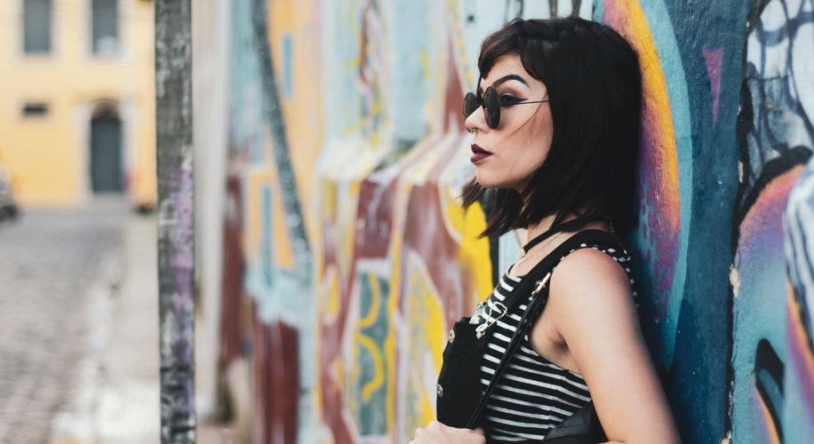 Woman with black hair wearing sunglasses, a striped shirt, and black overalls, leaning against a graffiti wall.