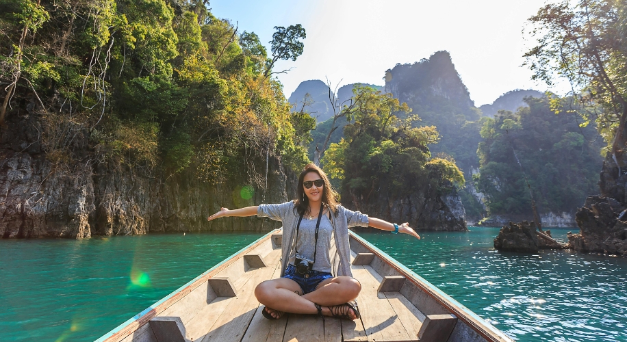 Person sitting cross-legged on a boat with arms outstretched, enjoying a scenic view of green water and forested mountains.