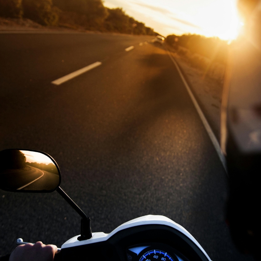 A person riding a scooter on an open road at sunset. The speedometer shows 0-80 km/h in blue.