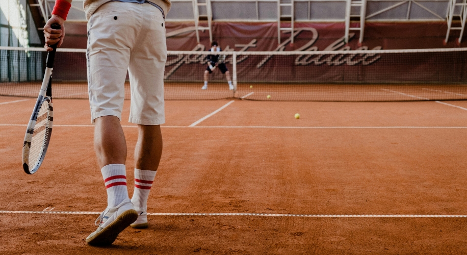 Tennis player holding a racket on a clay court, wearing white shorts and sneakers with red-striped socks.