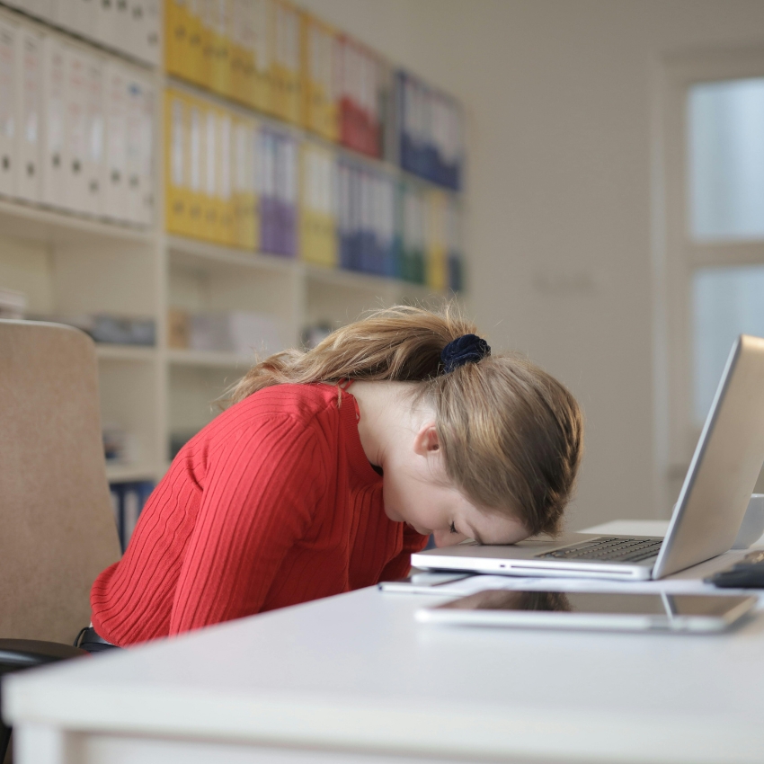 Person in a red sweater resting their head on a laptop screen, appearing exhausted. White desk in front.