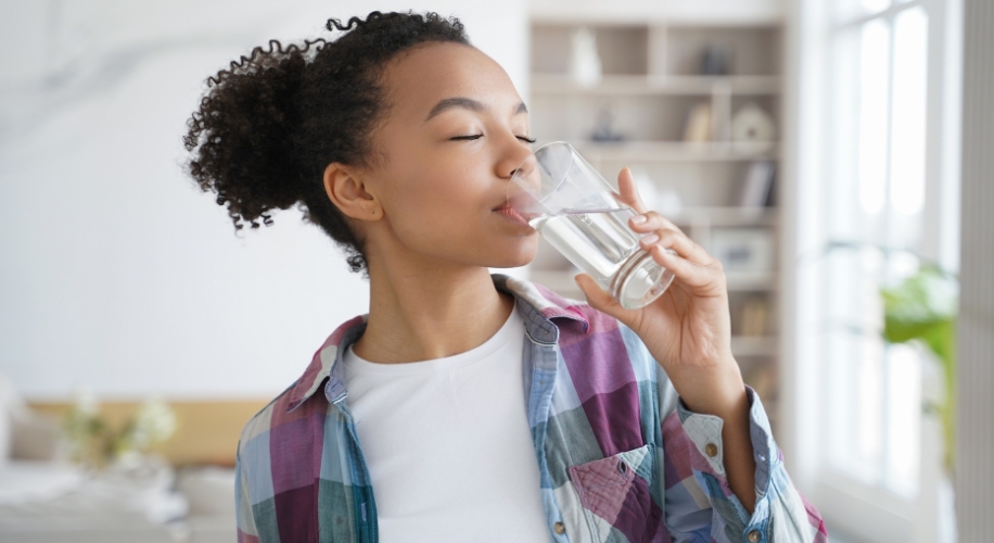 Woman drinking water from a glass, wearing a multicolored plaid shirt.