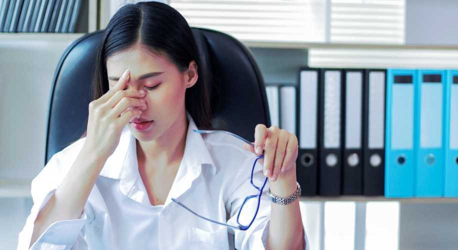 Woman in a white shirt, holding glasses, pinching her nose bridge, and appearing stressed.