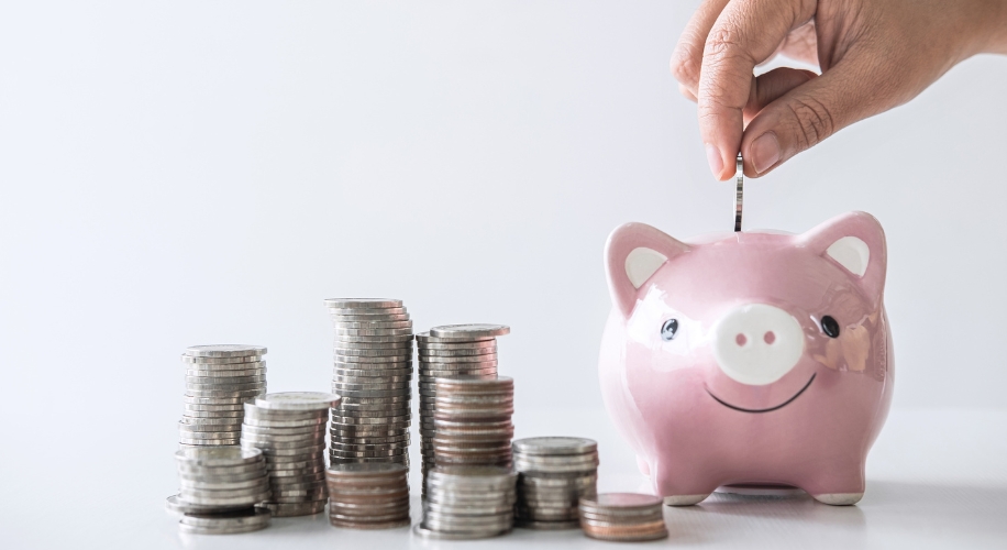 Hand placing coin into pink piggy bank next to stacks of coins.