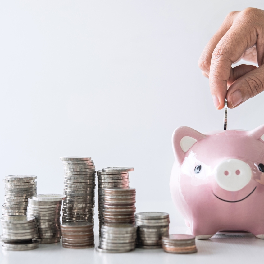 Hand placing a coin into a pink piggy bank next to multiple stacks of coins.