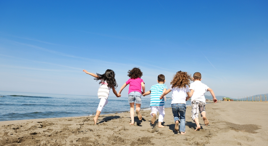 Children holding hands, running towards the ocean on a sandy beach. Clear blue sky overhead.