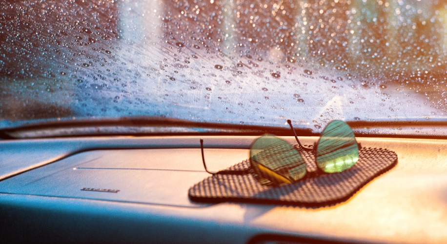 Green-tinted sunglasses on a black mat on a car dashboard with raindrops on the windshield.