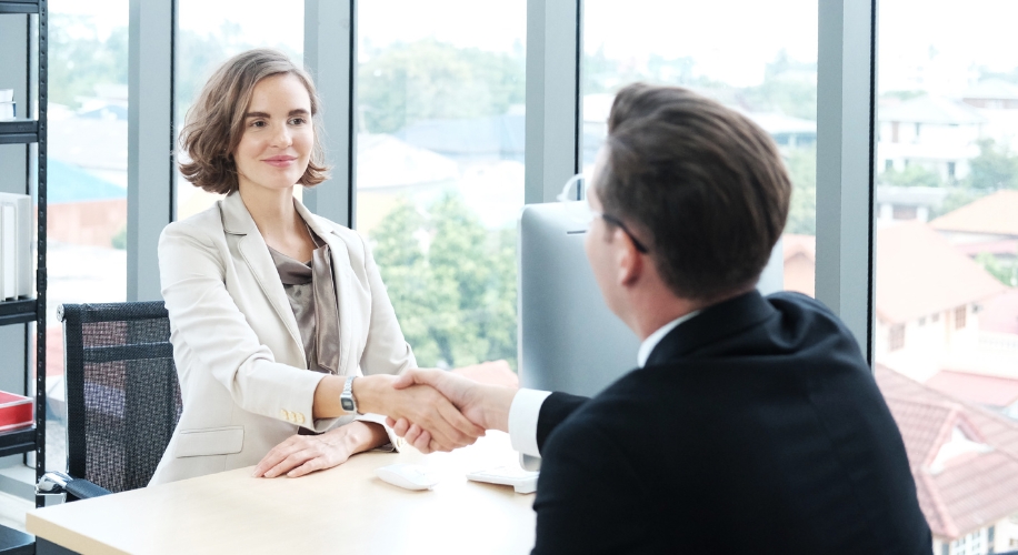 Two professionals shaking hands across a desk in a modern office setting.