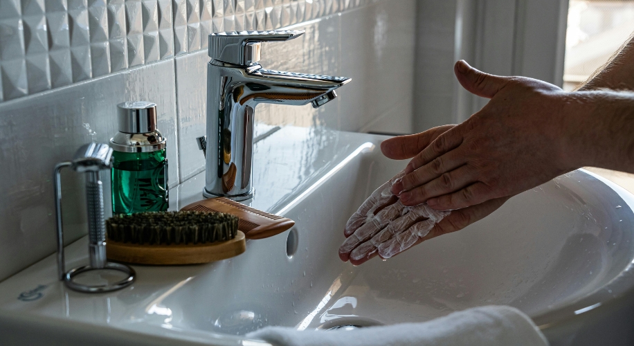 Hands washing with soap at a sink with a chrome faucet, next to a green bottle and a brush.