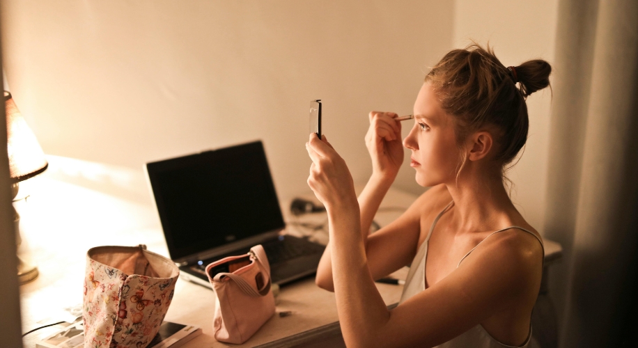 Woman applying makeup using a small mirror and brush at a desk. Nearby, open floral and pink pouches.