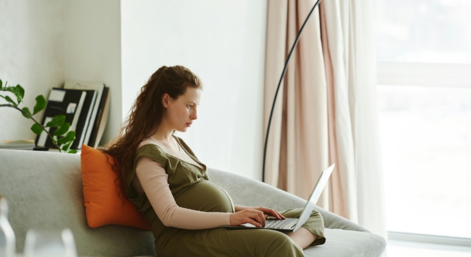 Pregnant woman sitting on a couch, working on a laptop with an orange pillow behind her back.