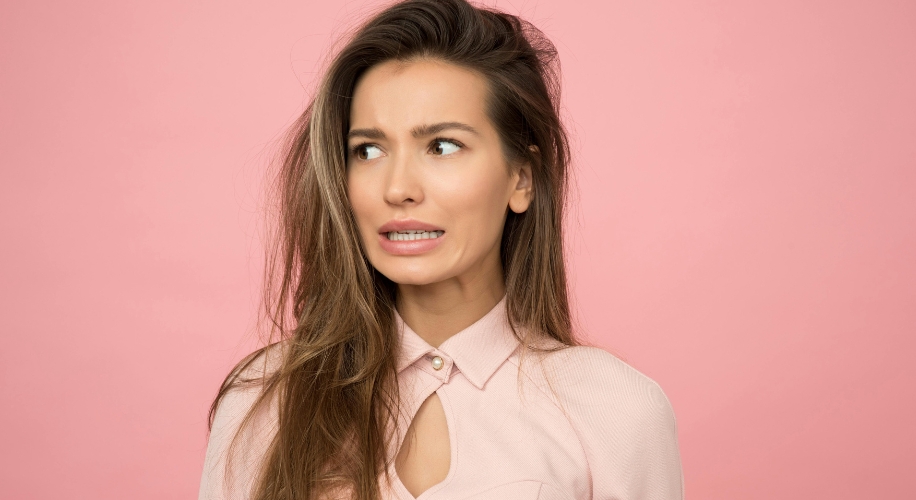 Woman wearing a light pink collared shirt with a nervous expression on a pink background.