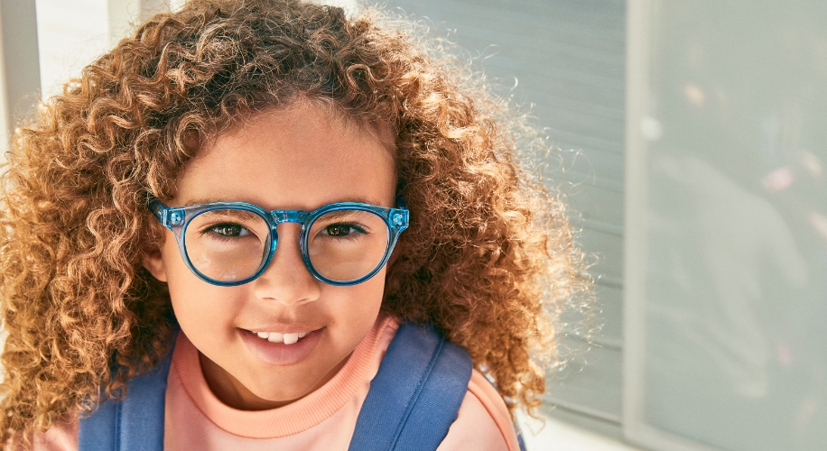 Child smiling and wearing blue eyeglasses with a blue backpack and a light pink shirt.