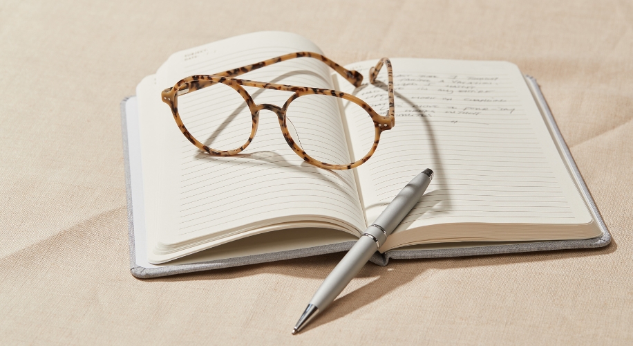 Open notebook with lined pages, tortoiseshell glasses, and a silver pen resting on a beige surface.