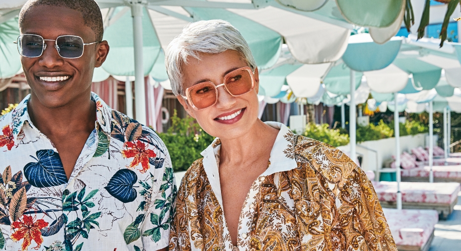 Two people wearing tropical print shirts and sunglasses, smiling outdoors under green and white umbrellas.