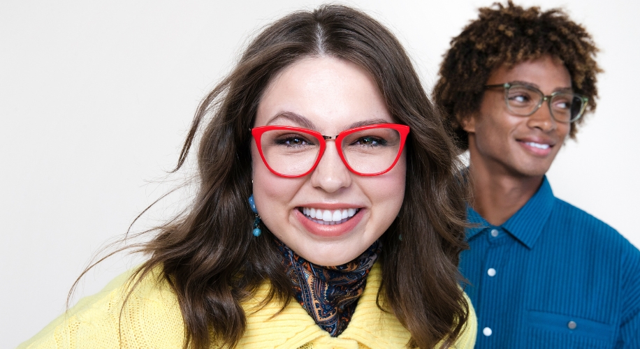 Woman wearing red cat-eye glasses, smiling directly at the camera. Another person in blue shirt, blurry in background.