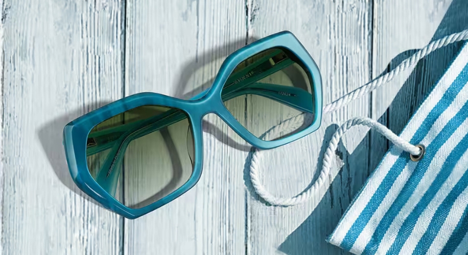 Large blue hexagonal sunglasses with green-tinted lenses, next to a blue and white striped cloth.