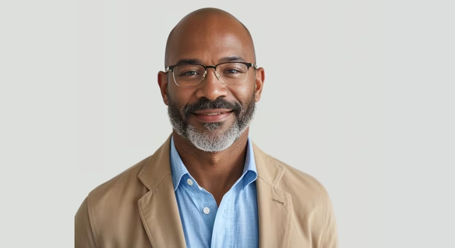 Man wearing glasses, a light blue shirt, and a beige blazer, smiling against a plain background.