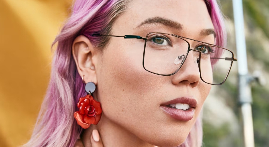 Woman wearing oversized rectangular glasses, large red rose earrings, and has pink hair.