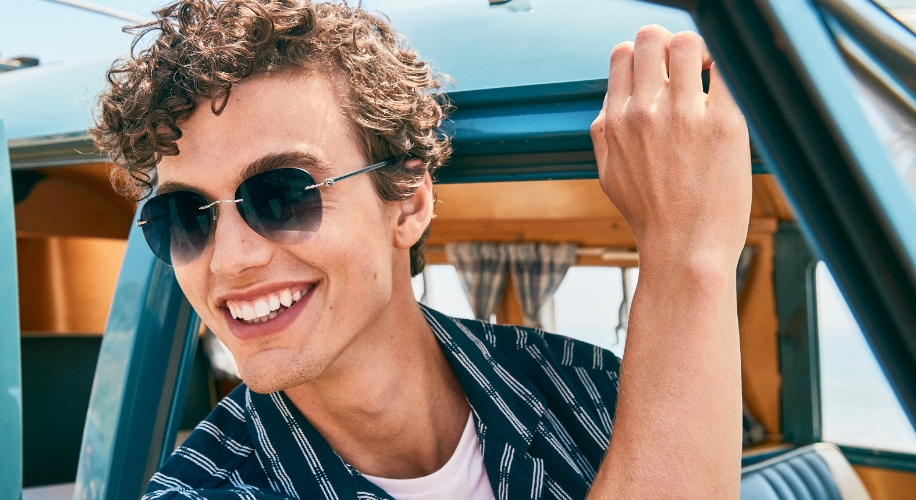 Man wearing round sunglasses and striped shirt, smiling while holding the top of a blue vehicle&#039;s door.