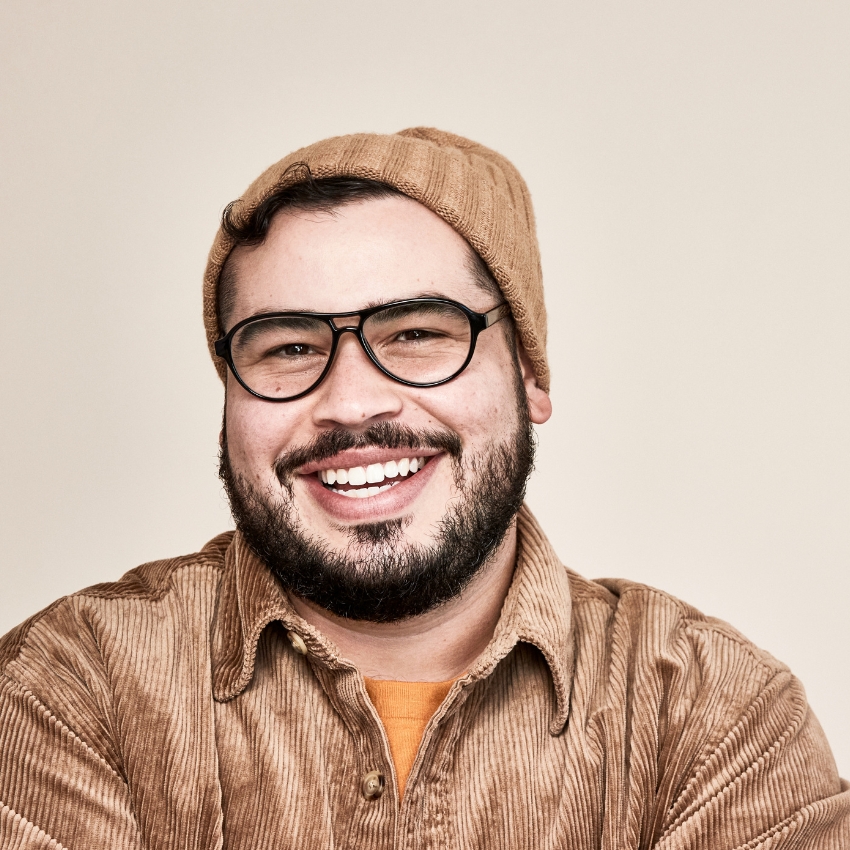 Man wearing glasses, a tan beanie, and a brown corduroy shirt, smiling.