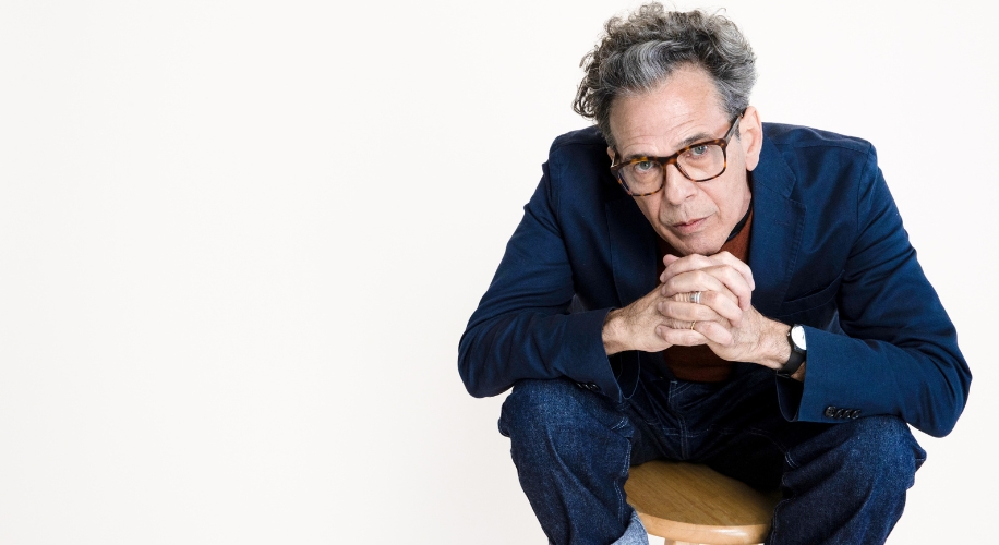 Man in glasses and a dark blazer sitting on a wooden stool, leaning forward with hands clasped.