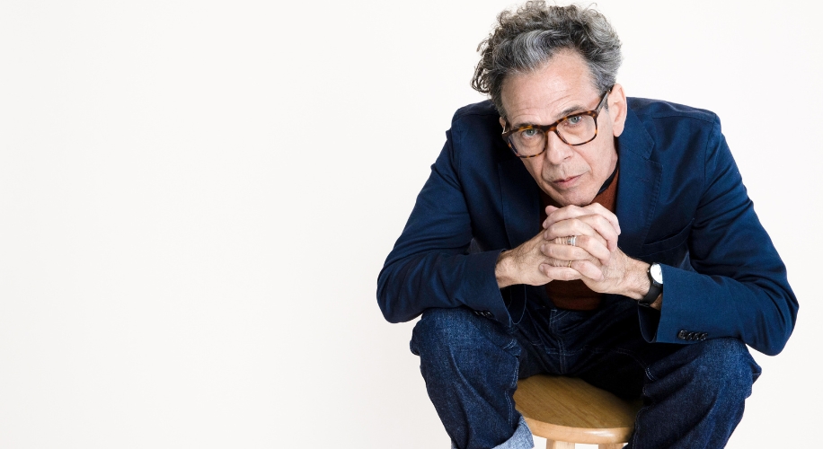 Man wearing glasses in blue blazer seated on a wooden stool with hands clasped and leaning forward.